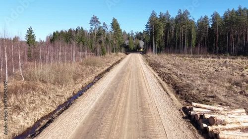 Dirt road leads through forested area towards a wooden house surrounded by tall trees, showcasing a clear blue sky and natural landscape in the background