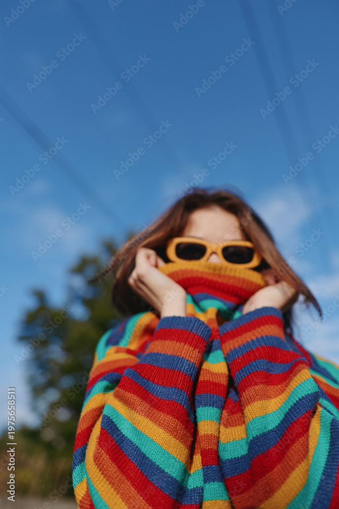 Fototapeta premium Young woman in a rainbow striped sweater and yellow sunglasses outdoors, playful pose pulling sweater over her lower face, casual outfit with blue sky background for lifestyle travel content.