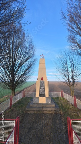 Memorial of liberation in Bolatice, Czech Republic, related to the Ostrava–Opava Offensive during World War II. The scene shows a rural field with a red star monument and a tree-lined avenue, symboliz