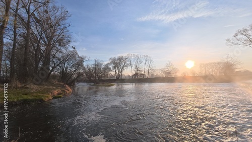 Small river weir with flowing water creating a gentle cascade. The scene captures movement, texture of water, and a calm natural atmosphere in a countryside setting.