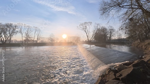 Morning weir on the Opava River at sunrise, with soft mist rising above the water and gentle light illuminating the peaceful river landscape. The scene captures calm morning nature atmosphere