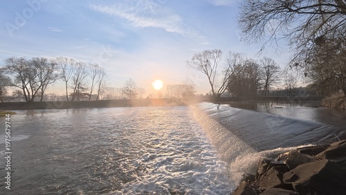 Morning weir on the Opava River at sunrise, with soft mist rising above the water and gentle light illuminating the peaceful river landscape. The scene captures calm morning nature atmosphere
