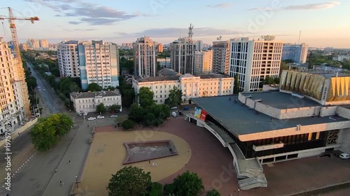 Odessa, Ukraine, August 15, 2025: City street view from above. Large multi-story buildings, square, green trees, asphalt road for cars, moving cars and walking people on sunny at sunset dawn