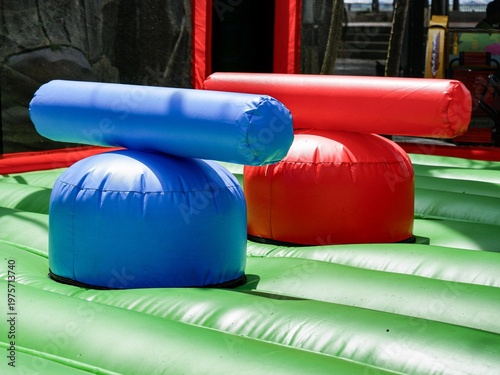 Colorful red and blue inflatable jousting pedestals on a green bouncy base. Perfect for competitive fun, outdoor events, and physical activity at children's amusement parks and carnivals.