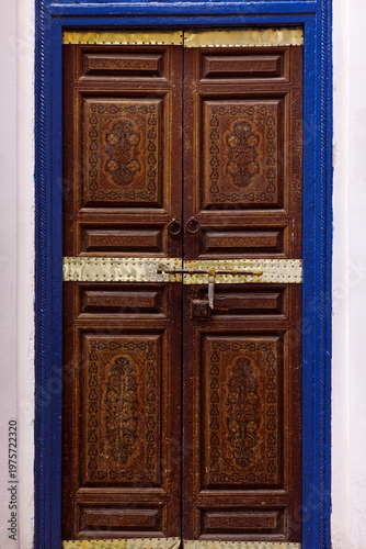Relief-carved, floral-motif painted old wood door with brass plates and blue frame, on a corner of the Bahia Palace Small Riad. Marrakesh-Morocco-263
