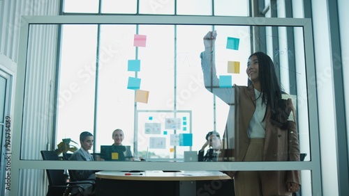 Foto Young Asian female company CEO explains marketing investment planning by writing on glass board, discusses, and brainstorms with multiracial partner team in meeting, business strategy presentation