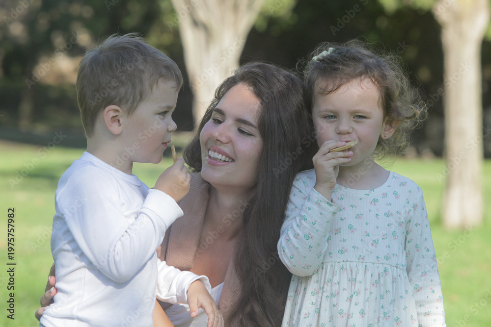 Fototapeta premium Mother and her kids share a happy moment together in a park having a snack, capturing natural bonding and connection in a summer outdoor portrait