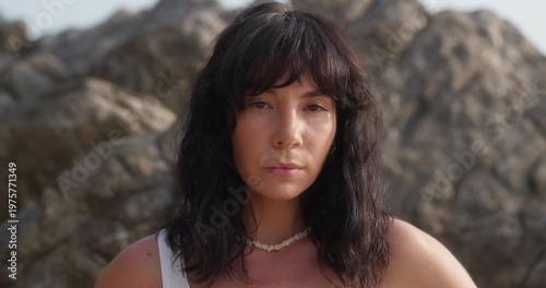 Portrait of long-haired brunette woman poses near rocks at exotic resort. Attractive lady with necklace stands near huge boulders closeup. Summertime fashion