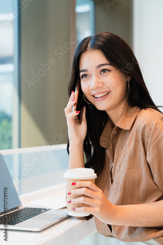 Young, happy, beautiful, professional Asian woman talking on smartphone, smiling, holding coffee cup in modern cafe.