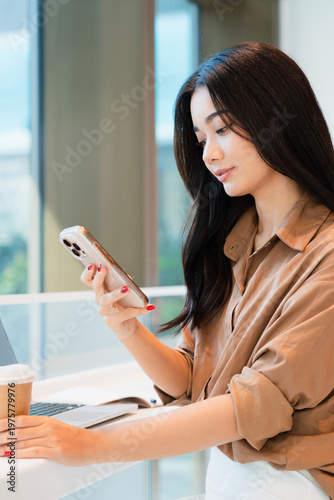 Young, confident, beautiful, professional Asian woman working at modern cafe, using, looking at smartphone.