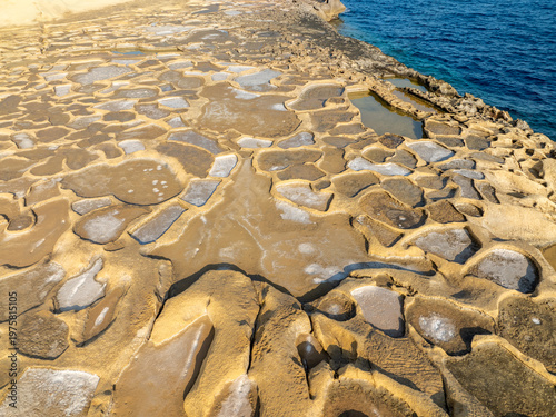 Aerial view of rugged, sun-baked salt pans shimmer with residual water, contrasting sharply with the deep blue sea, Iz-Zebbug, Gozo, Malta.
