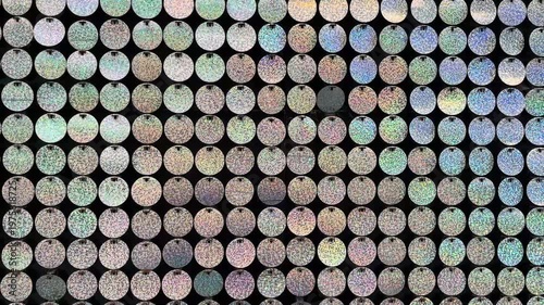 A close-up view of a textured surface covered with small, round, iridescent sequins in various colors. The background is dark, enhancing the shine of the sequins.