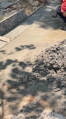 A man in a red shirt applies concrete to a surface. The scene includes a pile of wet concrete and tools for construction. Shadows are visible on the ground.