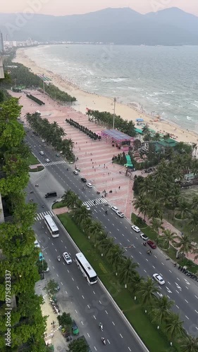 Aerial view of a beachside promenade with palm trees, a busy road, and people walking. The ocean waves crash against the shore under a sunset sky.