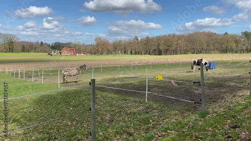 Wide shot of horses grazing in a fenced green pasture near a traditional farmhouse under a blue sky with fluffy white clouds on a sunny spring day.