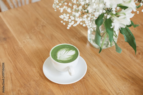 Overhead view of a white ceramic cup with matcha latte art and fresh white flowers in a glass vase on a wooden table.