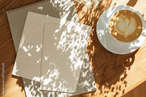 Aesthetic stationery mockup featuring a spiral notebook, blank cards a cup of cappuccino on a wooden table with sun shadows.