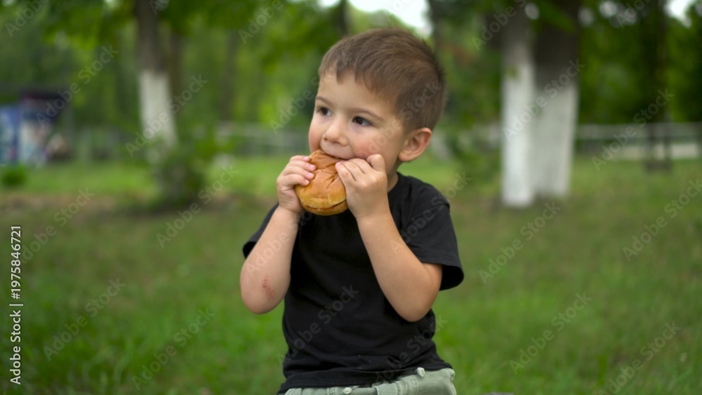 Fototapeta premium Three year old boy eats cheeseburger outdoors and looks at camera portrait 4k
