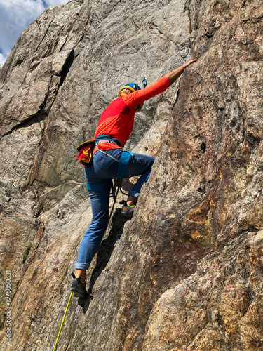 Woman wearing a safety helmet and bright orange long-sleeve shirt actively rock climbing a challenging granite rock face in Les Gaillands on a sunny day in the french alps, Chamonix.