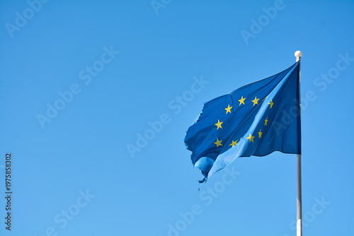 Damaged blue flag of the European Union featuring yellow stars under bright sunlight. Ideal for news coverage regarding international treaties and economic shifts.
