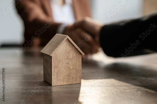 Wooden house model on a desk with a blurred handshake in the background. Real estate deal and property sale agreement between two business professionals. Mortgage approval and home ownership concept.