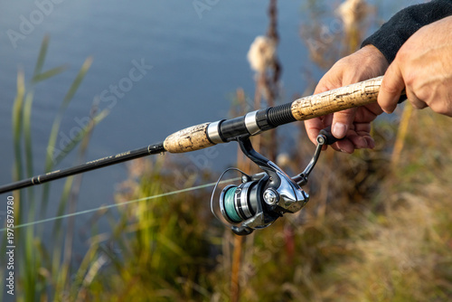 Canvas Print Hand holding fishing rod with spinning reel, angling gear by freshwater lake