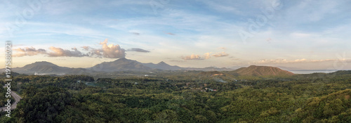 Aerial panorama from highway overpass captures Bakungan mountain range and river valley at sunset with golden light warming distant peaks and turquoise bay waters visible along the right horizon