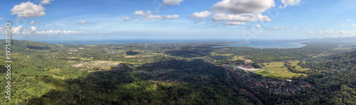 Wide aerial panorama from Irawan mountains shows the valley town spreading toward Honda Bay and open ocean with lush green hills in foreground and bright blue sky stretching across Palawan horizon