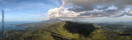 Wide aerial panorama reveals the full Mount Ari mountain range stretching across Bakungan district with lush green peaks under scattered clouds and ocean visible on the left horizon in Palawan