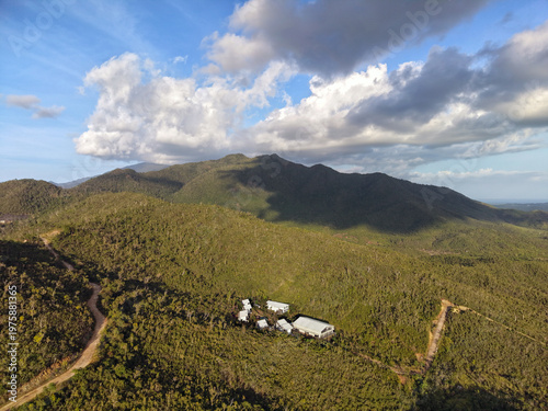 Aerial drone view shows rural compound buildings nestled at the base of Mount Ari surrounded by dense tropical forest with a winding dirt road cutting through lush green vegetation in Bakungan Palawan