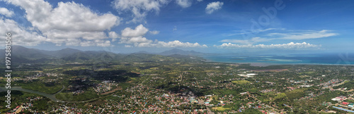Ultra-wide aerial panorama captures Puerto Princesa cityscape spreading from mountain foothills to turquoise bay waters with cumulus clouds dotting the blue sky above the Palawan island landscape
