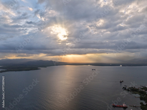 Aerial drone view captures golden sunset rays breaking through heavy clouds above Puerto Princesa Harbor with cargo ships anchored in calm bay waters and mountainous Palawan shoreline beyond