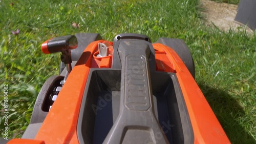 Closeup POV shot over the front of a modern battery operated, cordless, garden or yard mower, which is being pushed forward while cutting a grass lawn on a warm sunny day.