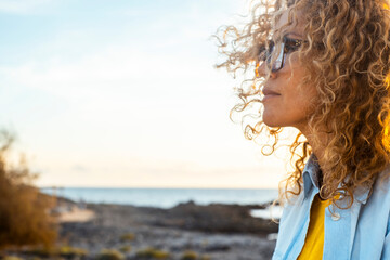 Thoughtful woman in profile at seaside during golden hour sunset with curly hair and glasses