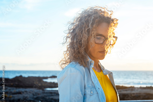 Fotografie Thoughtful mature woman in glasses standing by the beach at sunset in casual coa