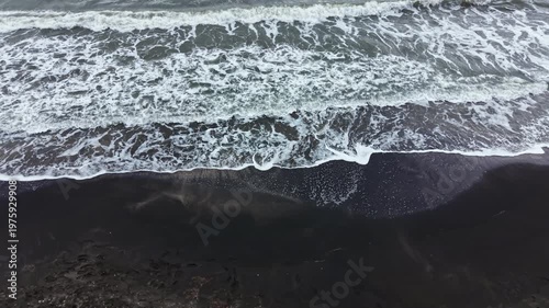 Waves move back and forth on a black sand beach, churning up foam. The sky is cloudy, and the wind blows strongly over the ocean. Water splashes as the tide comes in and goes out.