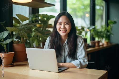 Free Download Asian woman smiling at camera while working on laptop in a green home office - GFXLoad
