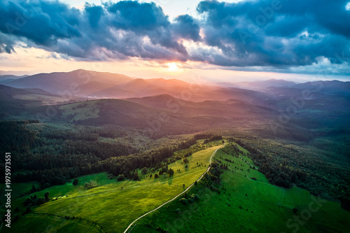 Aerial view breathtaking sunset over forest. Sun's rays break through dramatic clouds, casting golden glow across rolling hills and valleys. Distant mountains creating serene and picturesque scene.
