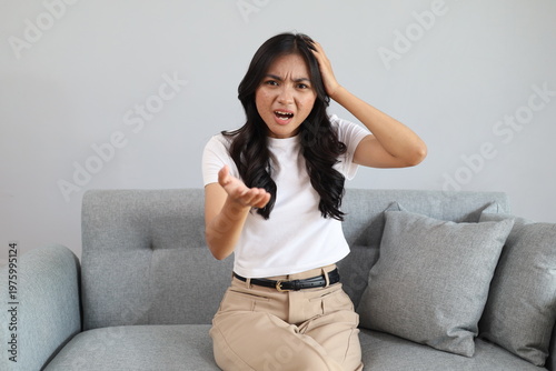 Upset young Asian woman arguing and talking while sitting on the sofa in her living room, angrily complaining about various problems while looking at the camera.