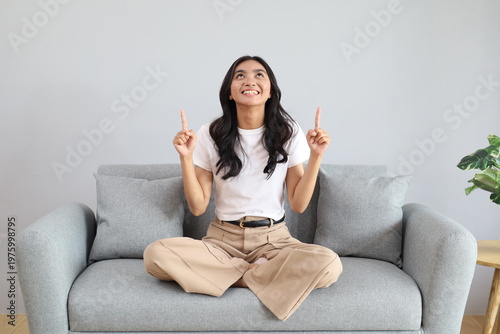 Billede på lærred A smiling young Asian woman looks and points up at the empty space for your ad text, sitting cross-legged on the sofa in the living room of the house