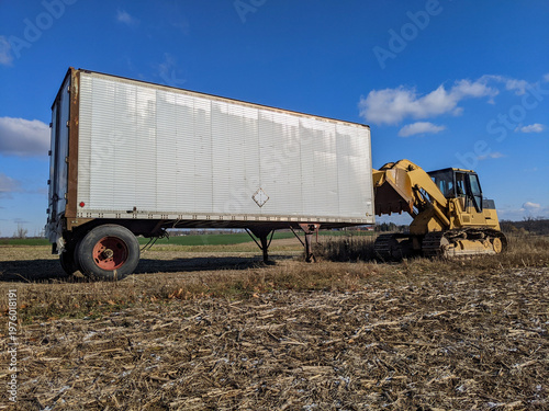 A yellow excavator pulls an old trailer on a harvested rural field, captured under a bright blue sky with soft clouds. The scene evokes agriculture, heavy equipment, transportation and outdoor work.