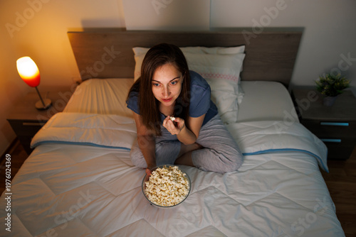 Woman eating popcorn while watching funny movie on TV in bed at home at night