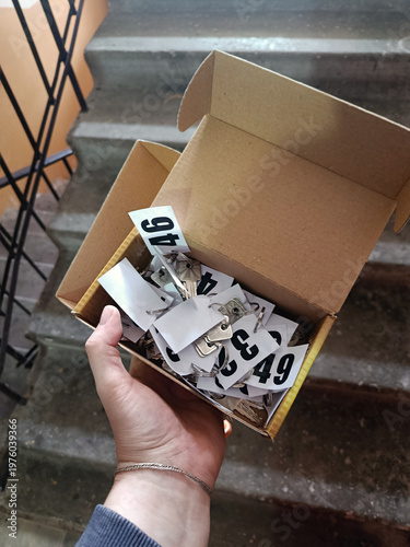 A box with small keys with paper numbers written on them. A box with keys in a hand.