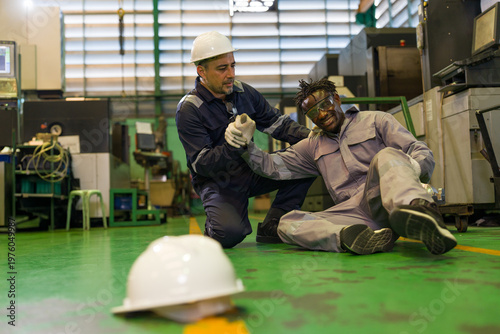 Injured worker lying on floor after industrial accident in factory with colleague providing first aid. Supervisor helping hurt technician after falling down in manufacturing factory floor safety