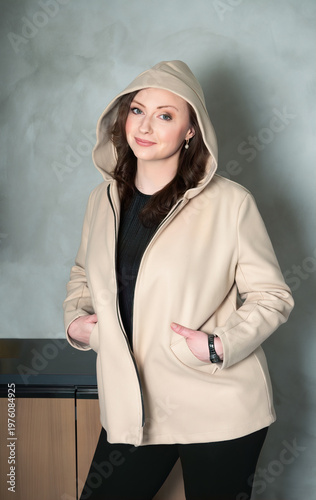 Confident young woman with long wavy brown hair wearing a stylish beige hooded jacket with hands in pockets looking at camera with a gentle smile while posing in a modern minimalist office interior