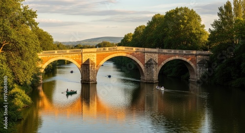 A picturesque stone bridge with arches spanning a river, surrounded by lush green trees and a clear blue sky with a few scattered clouds. 