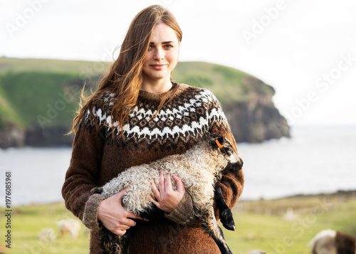 A girl wearing an Icelandic sweater is standing in Iceland holding a young lamb in her arms.