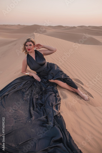Beautiful woman sitting on sand dunes in elegant black dress.