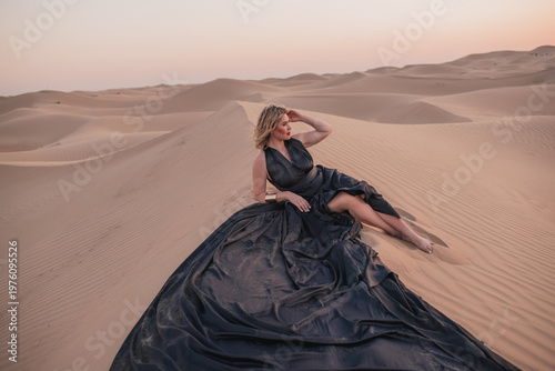 Beautiful woman sitting on sand dunes in elegant black dress.