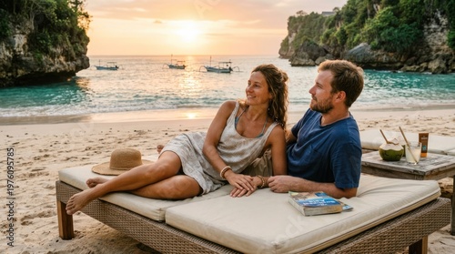A happy couple relaxes on a lounge bed overlooking a beautiful tropical beach at sunset. A woman and a man are sitting side-by-side, enjoying the view and a drink.